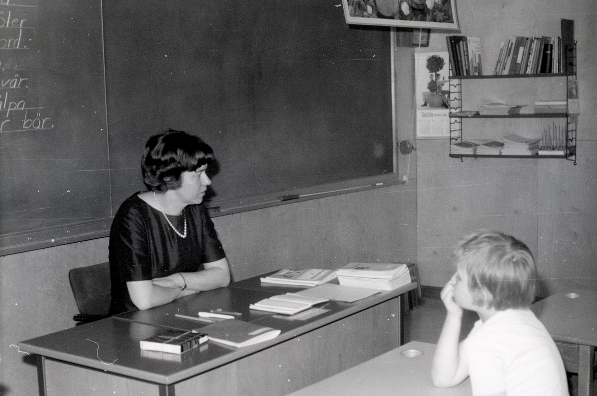 Lärare Ruth Singmyr sittandes vid en kateder i Kålleredsskolan (senare Brattåsskolan) cirka 1965. Bakom henne ses "svarta tavlan". Hon arbetade i Kållered från 1964 -. Okänd elev sitter i bänken framför henne.