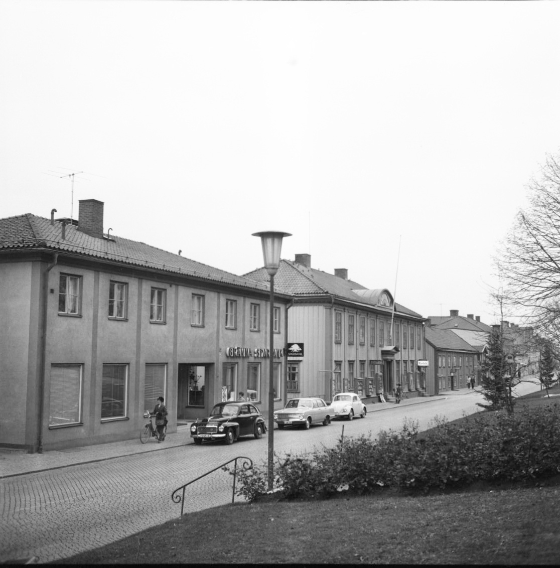 Brahegatan med Sparbankshuset och Hallska gården. Bilar står parkerade utmed fastigheterna. En kvinna leder en cykel söderut på trottoaren.