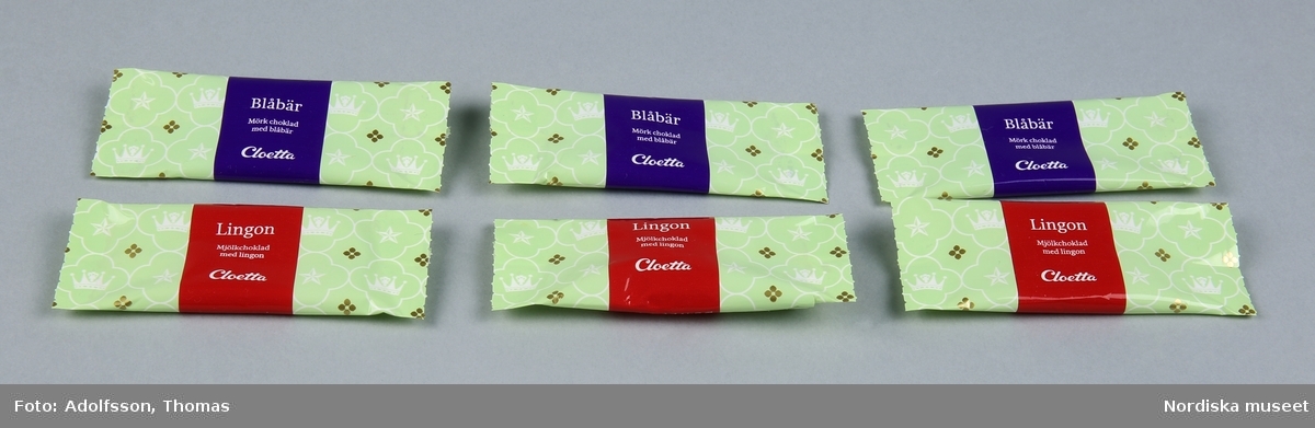 a-f) 6 st plastomslag för chokladbitar. Grön bottenfärg med samma mönster som asken (kronor och Nordstjärnan inskrivna i fyrpass på grön botten).  På tre av dem lila tvärband med text "Blåbär / Mörk choklad med blåbär / Cloetta / 10 g" samt symboler för att plasten kan återvinnas. På tre av dem rött tvärband "Lingon / Mjölkchoklad med Lingon / Cloetta / 10 g" samt symboler för att plasten kan återvinnas. 
/Leif Wallin 2015-01-12