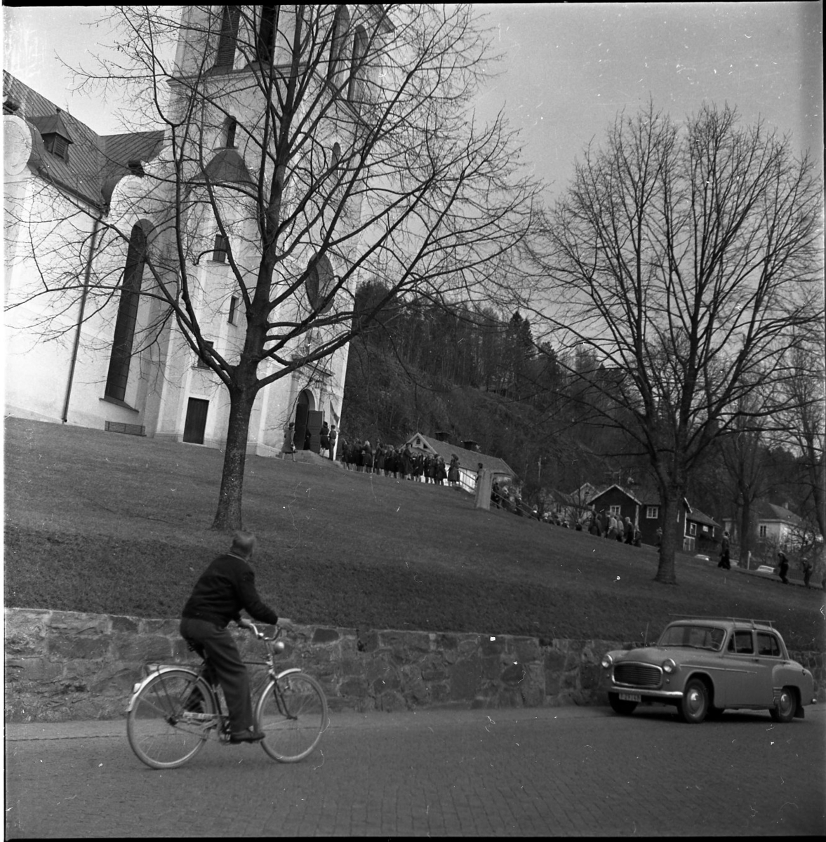 Ett led med ungdomar på väg in i Gränna kyrka. Det är troligen scoutupptagning. En cyklist på väg söderut och en bil står parkerad norrut nedanför kyrkmuren.