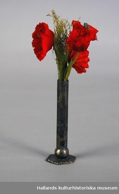Dockskåpsvas med blommor. Nysilvervas, sex-kantig, längst ned en kulformad del och en sex-kantig fot. Tygblommor i grönt och rött. Blommornas längd: 10 cm.