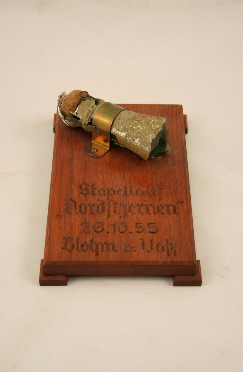 Ei treplate påmontert den avslåtte tuten fra ei champagneflaske. I plata er det innskåret tekst: "Stapellauf "Nordstjernen" 26.10.55 Blohm & Voß".