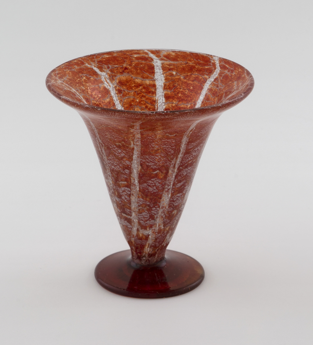 Konisk vase i Ikorakrystall som hviler på en sirkulær fot. Korpus har en marmoreringslignede overflate, hvor melkehvite og klare partier bryter opp den rødfargede bunnen.