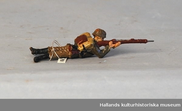 Leksakssoldat av trämassa på ståltrådsskelett i tysk uniform. Soldaten ligger raklång och skjuter med sitt gevär. Målad i grönt, brunt, svart, gult och skärt.
