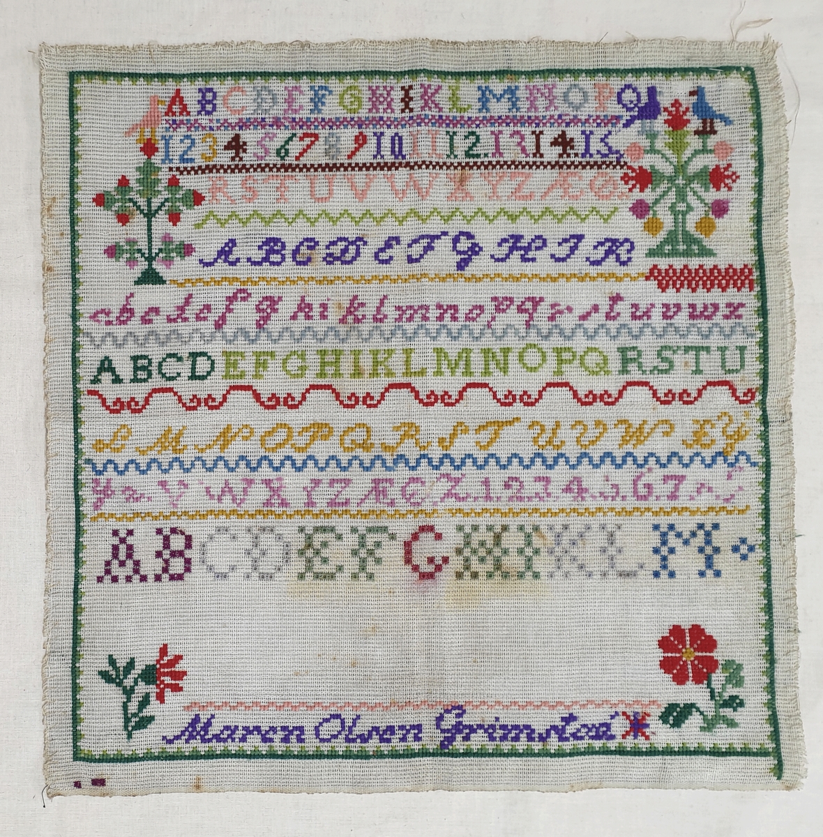 Brodert navneduk med bokstaver og tall i forskjellige farger på strie (kanvas).