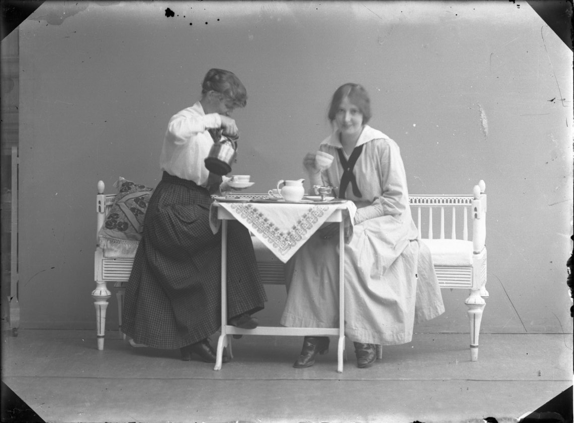 Ateljébild med syskonen Karin och Anna-Lisa Rythén som sitter i en soffa vid ett dukat bord med kaffe. Karin häller upp i sin kopp och Anna-Lisa håller en kopp i sin hand.