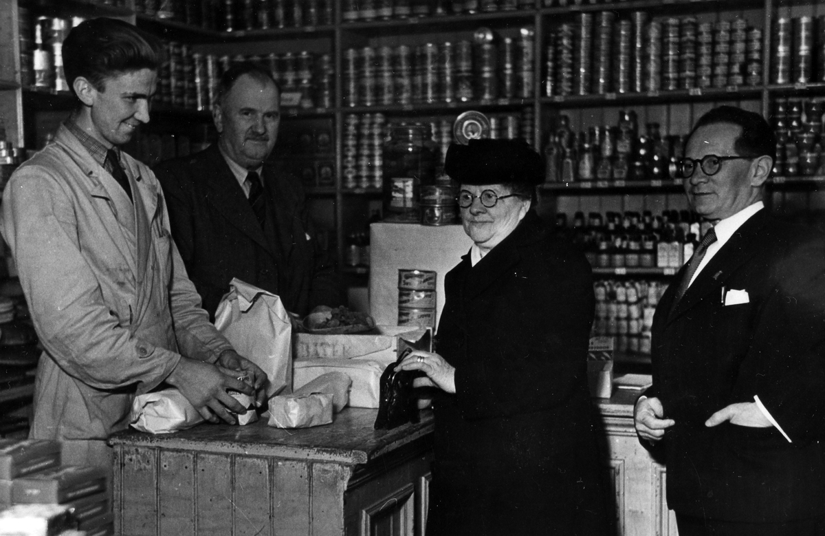 I Claes Nygrens Speceriaffär står Claes Nygren och Sigge Gisleskog bakom speceridisken och betjänar två kunder, Lena Karlsson och en okänd man. Hyllorna bakom fyllda med konserver.
