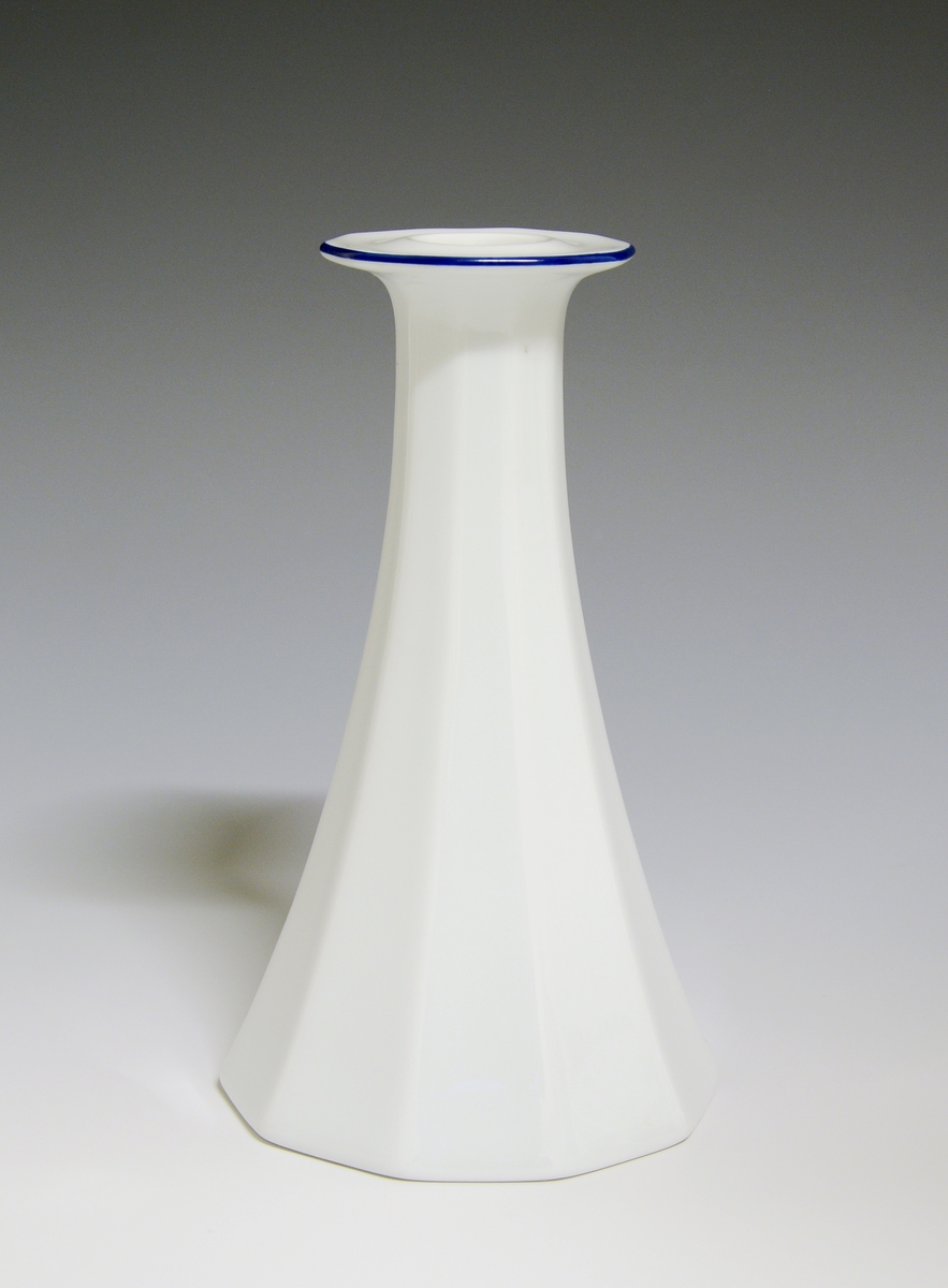 Mangekantet lysestake av porselen med hvit glasur, dekorert med en enkel blå strek langs lysestakens krage.
Modell: Octavia, tegnet av Grete Rønning i 1977
Dekor: Blå strek