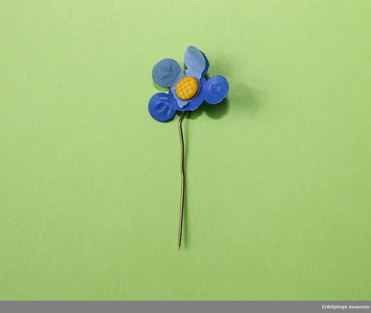 Majblomma från 1937.
Blomman är gjord av ljusblå och mörkblå celluloid. Blomman har två övre blad i ljusblått (tre saknas) och fyra undre blad i mörkblått (en saknas) samt en gul mittknapp, även denna gjord av celluloid. 
Det som håller blomman samman är en nål av mässing.