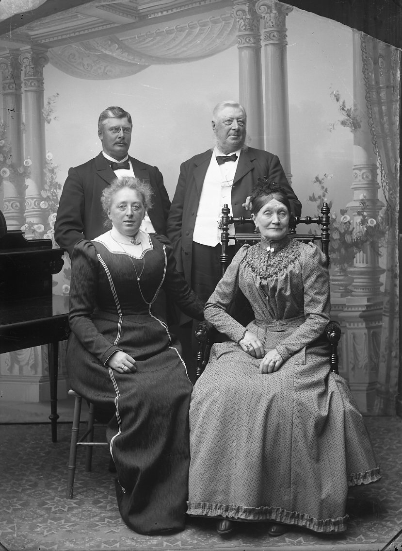 Två par i en fotoateljé. Kvinnan till vänster sitter på en stol och vilar vänster arm på ett skrivbord.  Kvinnan till höger sitter i en gungstol och vilar fötterna på en kudde. Kvinnornas män står bakom dem. Det yngre paret, till vänster, är grosshandlare Mårten Persson och Anna Granlund.