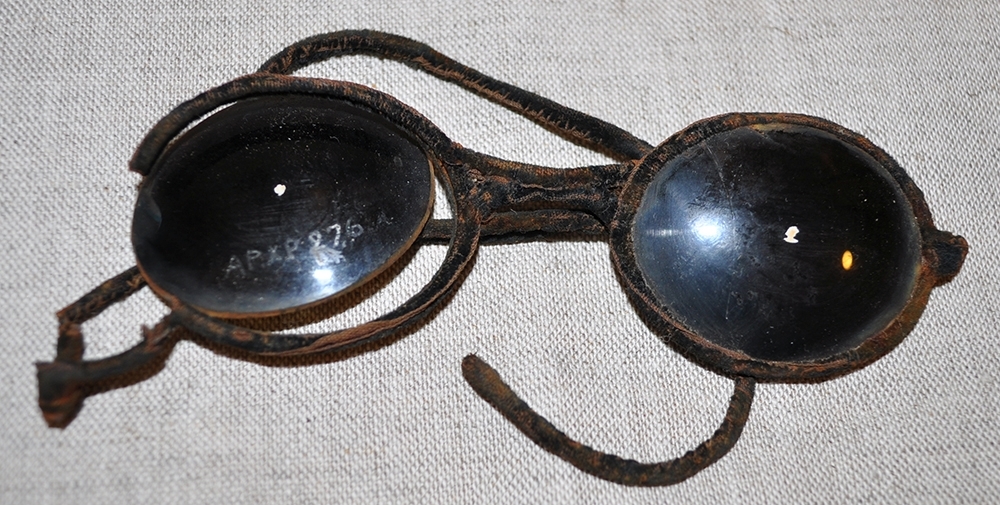 Glasögon i ett fodral. A: Svarta solglasögon, ovala glas. Ett glas är löst. Trasiga skalmar. B: Fodral i papper som består av tre dela som skjuts ihop.