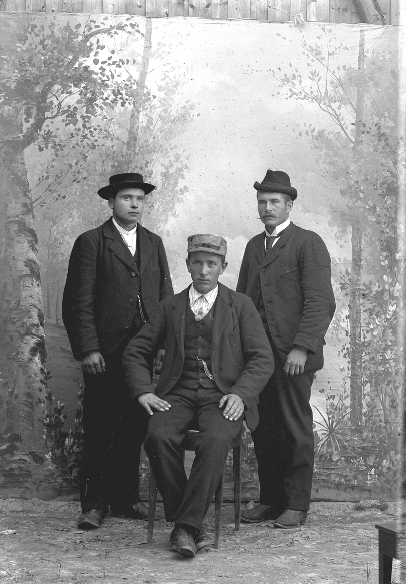 Tre unge menn, en sitter (har lys hatt/lue) og to står (mørke hatter). Alle kledd i mørke dresser.