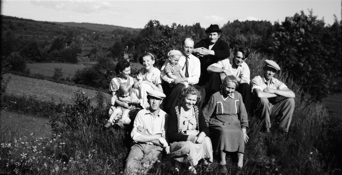 En grupp människor, åtta vuxna och tre barn, sitter på en gräskulle en solig dag. Längst fram till höger sitter Anna i Planterhagen och mannen i hatt i mitten är hennes man Herman.
(Se även bild EA0484)