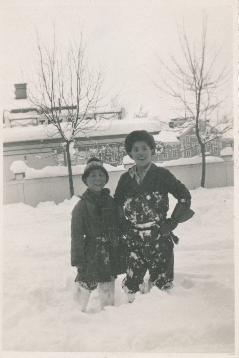 Två pojkar står i snön och ler mot kameran. De har troligen lek i snön då deras kläder är snöiga. I bakgrunden skymtar en bensinstation.