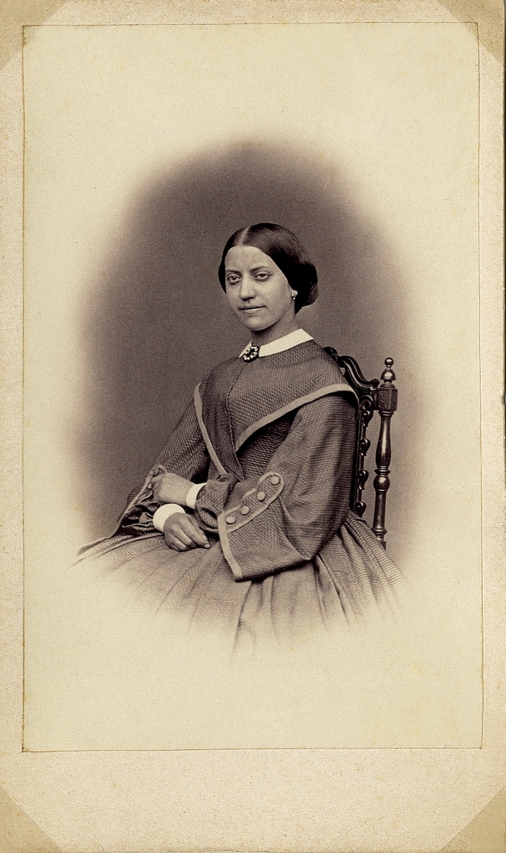 Porträttfoto av en kvinna i krinolin med liten vit krage. Vid kragen syns en brosch. Hon sitter på en högryggad stol. 
Knäbild, halvprofil. Ateljéfoto.