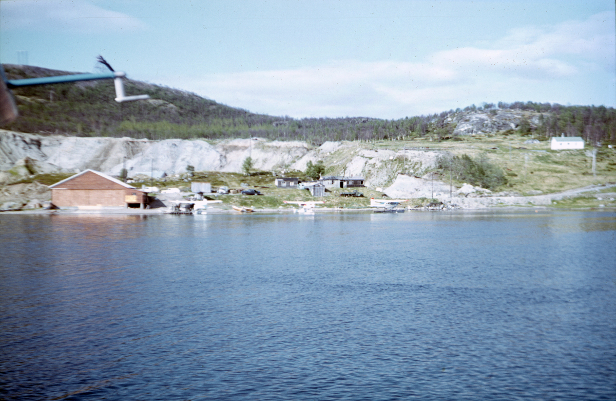 Hovedbasen til Varangerfly med sjøflyhangaren på Sandnes, Langfjorden i Sør-Varanger.Tre sjøfly ligger fortøyd i blåser nær stranden. På land ligger ei større rød brygge og flere mindre buer.
