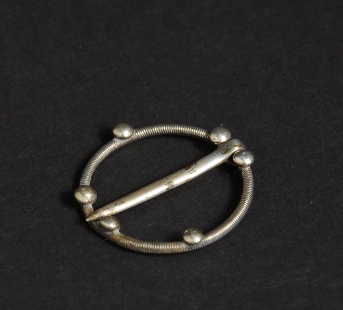 Spenne av sølv. Spennen har sirkulær form med dekorative tverrlinjer, og seks runde kuler.