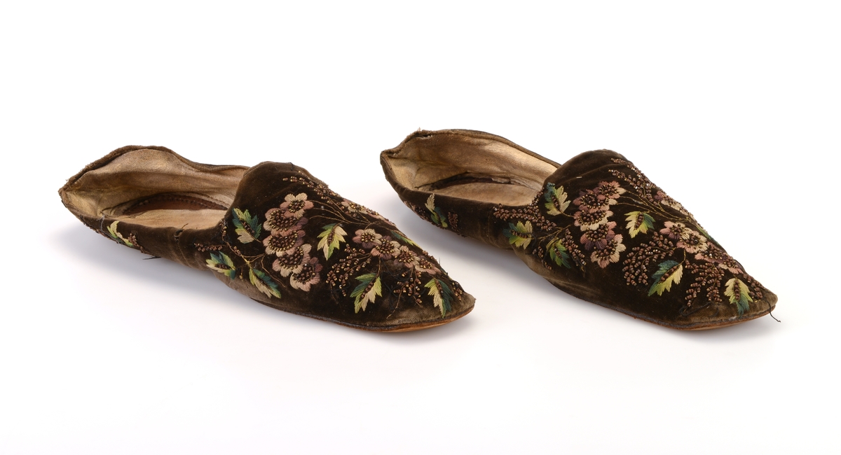 Et par damesko/tøfler sydd av brunsvart fløyel. På fløyelen er det brodert blomster og blader i ulike nyanser av lilla og grønt samt gråhvitt. Det er brodert med plattsøm og attersting. Det er også brodert på rikelig med metallperler som en gang har vært sølvfarget, men som nå er brun. Broderiene er både foran og bak på skoene
Innvendig er skoene foret med ubleket linstoff i den fremre delen av skoen. Den bakre delen er foret med skinn som en gang har vært hvit. Innersålen er av hvitt skinn. Yttersålen er av brunt lær. Skoene er firkantet foran og er helt flat.