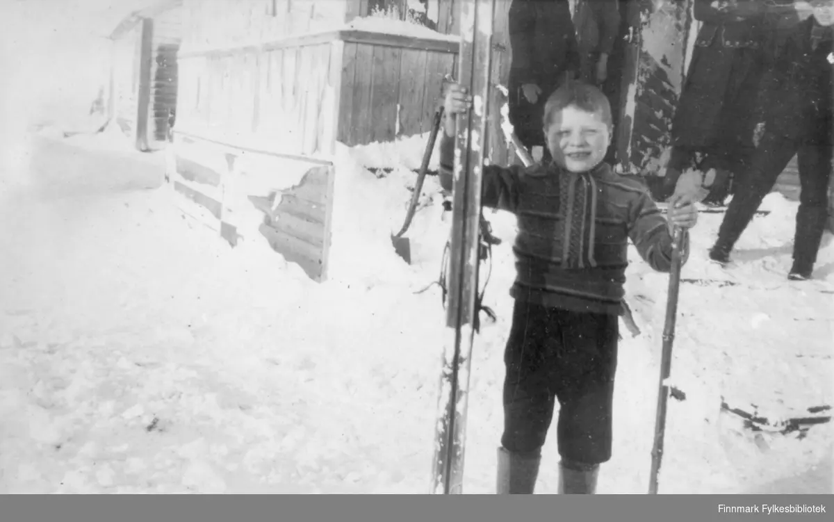 En gutt holder ski i hånden. Mye snø på bakken/veien. Noen personer står i bakgrunn ved døråpningen til huset.