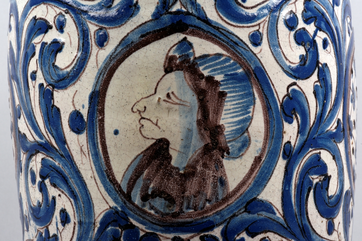 Vitt krus med två öron och en smal hals, tillverkat av fajans. Dekorerad med blått, runt livet fyra medaljonger, en med ett gumhuvud och på motstående sida en medaljong med inskription AQVA CICOILA 1773.