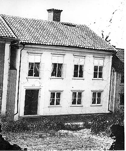 Tvåvåningshus med putsad fasad troligen motsvarande dagens Brahegatan 63 i Gränna. I två öppna fönster på andra våningen syns en man respektive en kvinna med en hund. Det kan vara fastighetens ägare fd översten Carl Vilhelm Ahlgren respektive Amalia Sofia Crona.