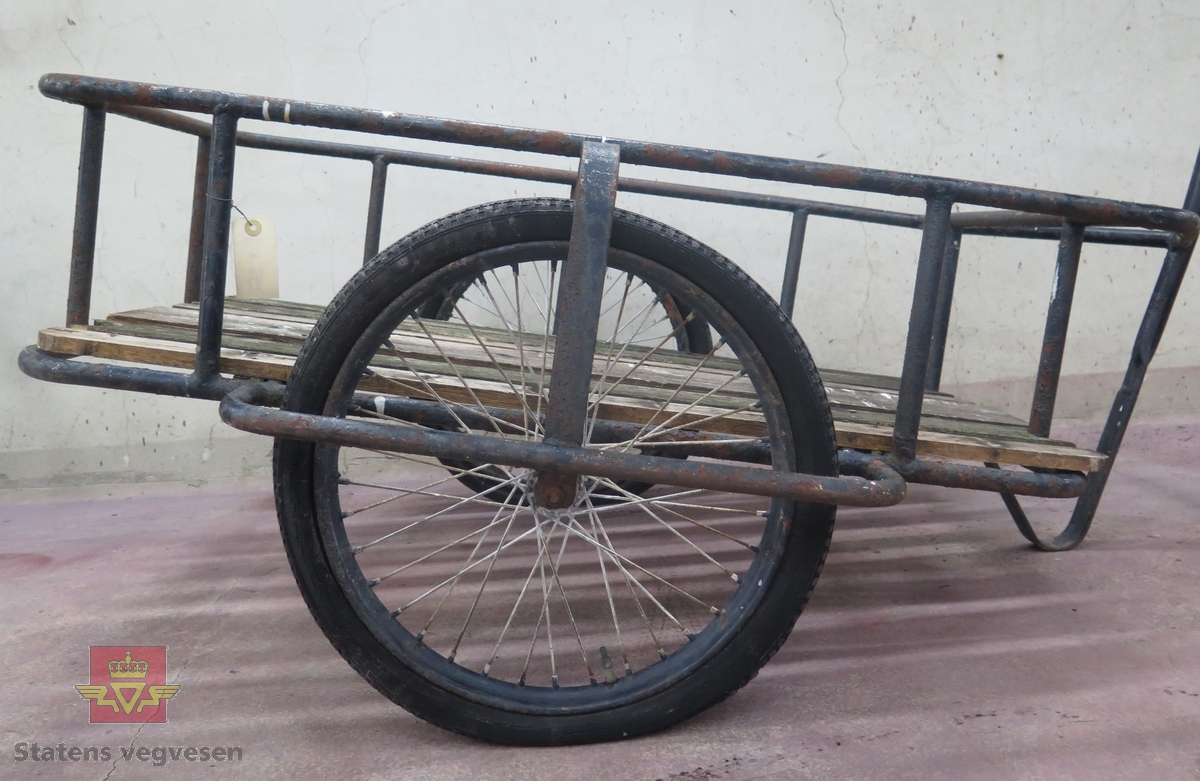 Tohjuls tilhenger for transport etter sykkel, Svart ramme og kontruksjon. Gulv av tre, har rester av grønn farge. Preget av rust.