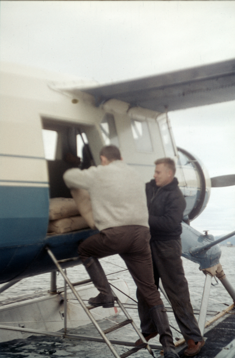 Nor-Wings utfører et transportoppdrag, trolig i forbindelse med vannkraftutbygging på Helgeland. To flygere laster sement ombord i en Norsman som ligger fortøyd til ei flytebrygge ved kysten.