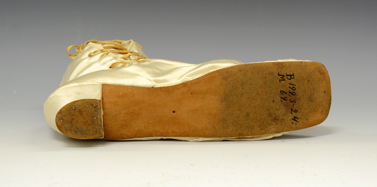 Silkestøvler. Hvite silkestøvler med høyt skaft og snøring. Sløyfe på tå og liten hæl. Silkestøvlene tilhører empirkjole TGM-BM.1923-24:068.