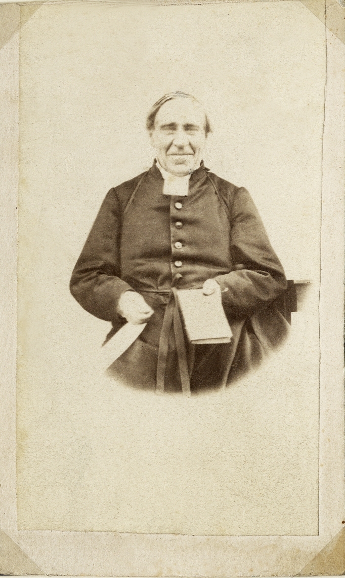 Porträttfoto av en äldre man i prästdräkt m.m. 
Midjebild, en face. Ateljéfoto.