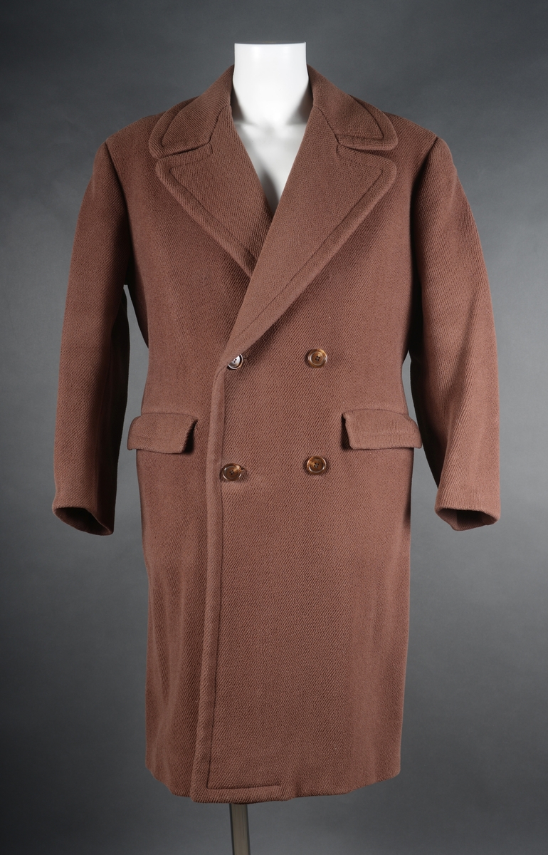 Frakk av tykk ull, med paspoilerte lommer og spensel bak. 4 knapper foran.