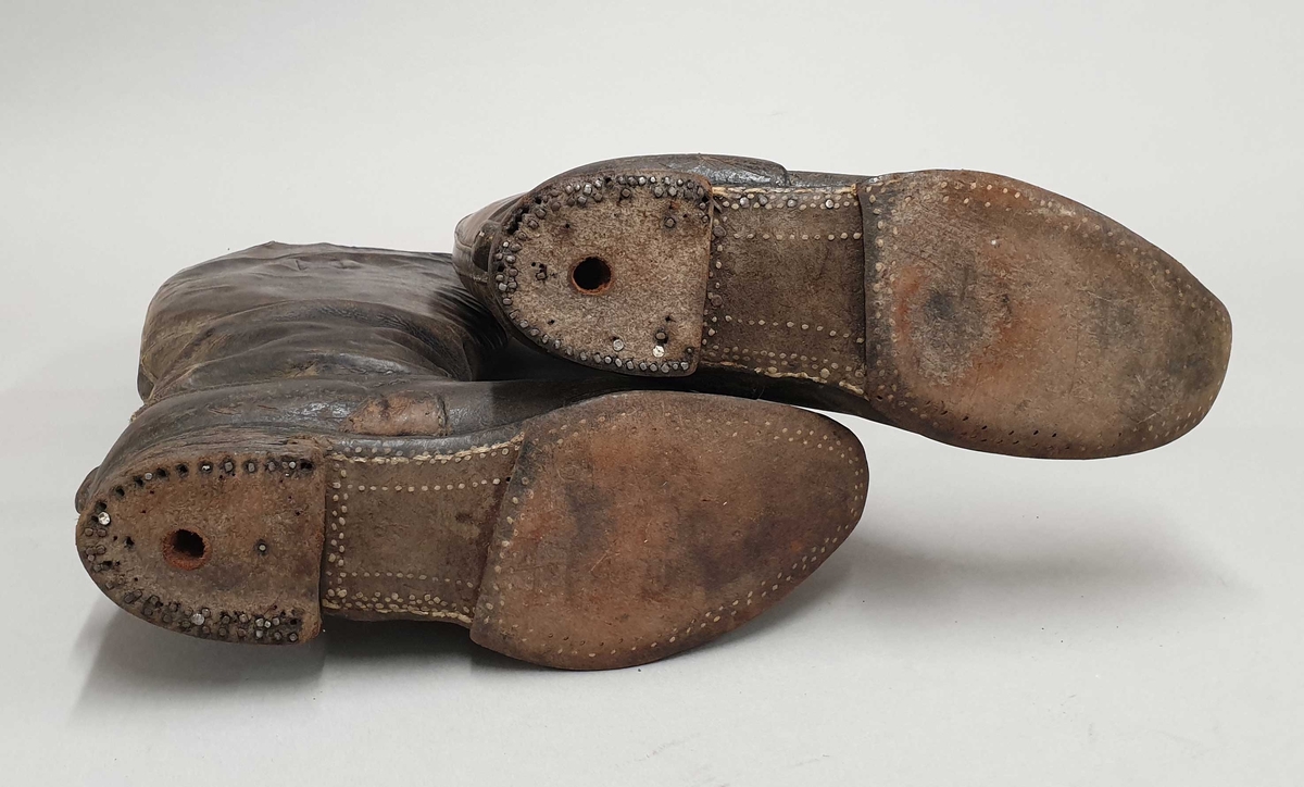 Ett par høye støvler av lær, med hemper øverst. Nagler på undersiden av sålen, og hull i begge hælene.