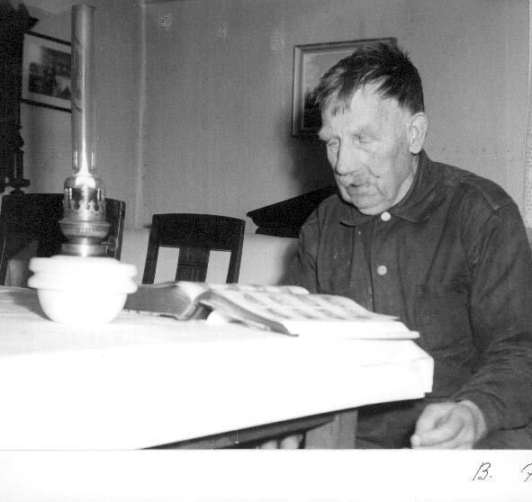 En gammal man, Karl Alfred Johansson Rehn i Sonaby, sitter vid ett bord med en tänd fotogenlampa och uppslagen bok framför sig.