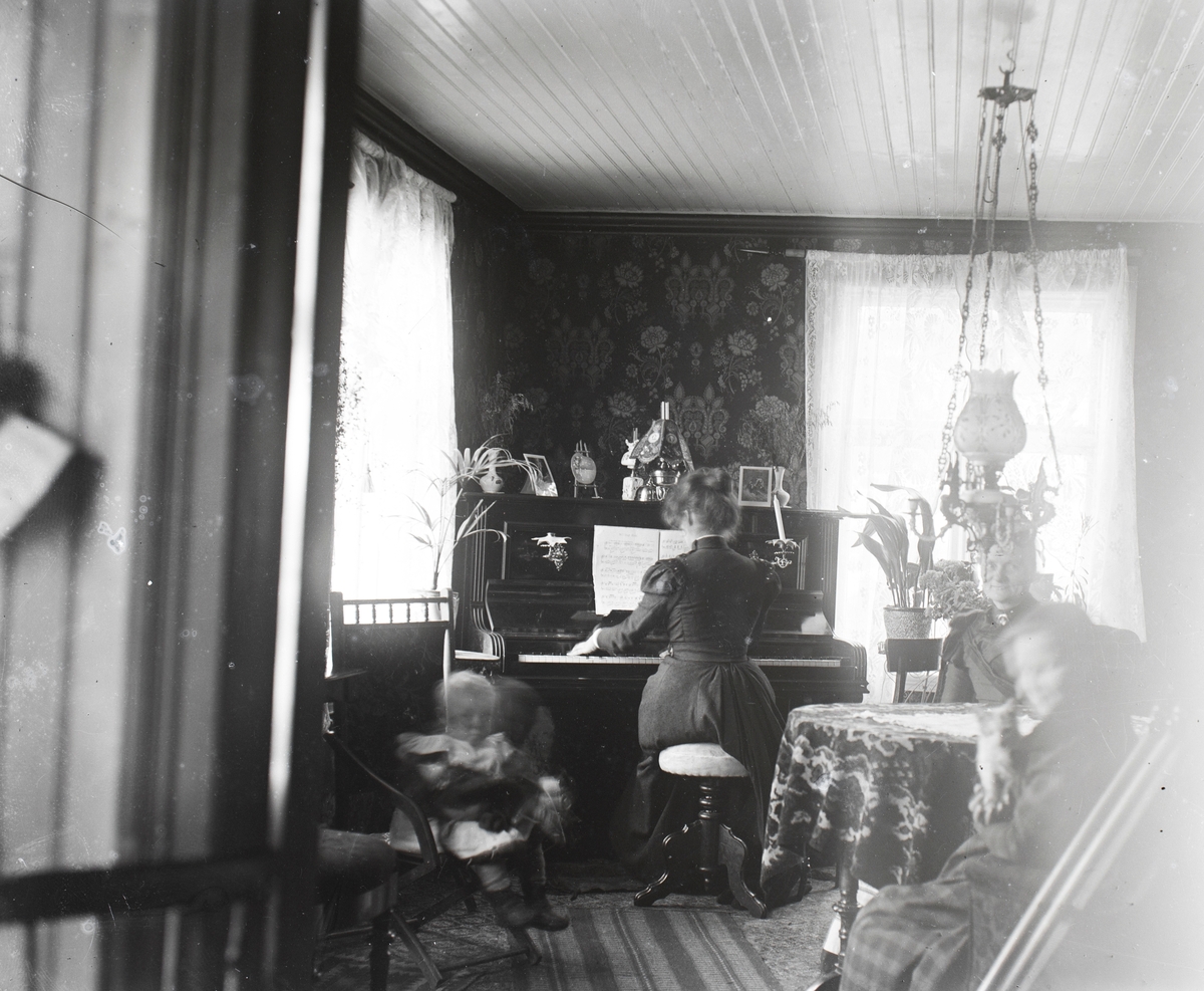 Motiv av familien Aston i stuen. Ved pianoet sitter Evelyn Aston.