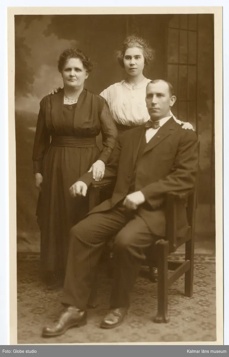 Familjeporträtt? Till vänster står en mörkhårig kvinna klädd i svart klänning.Till höger om henne står en yngre ljushårig kvinna, klädd i ljus blus. Längst till höger sitter en man på en stol, han är klädd i mörk kostym.