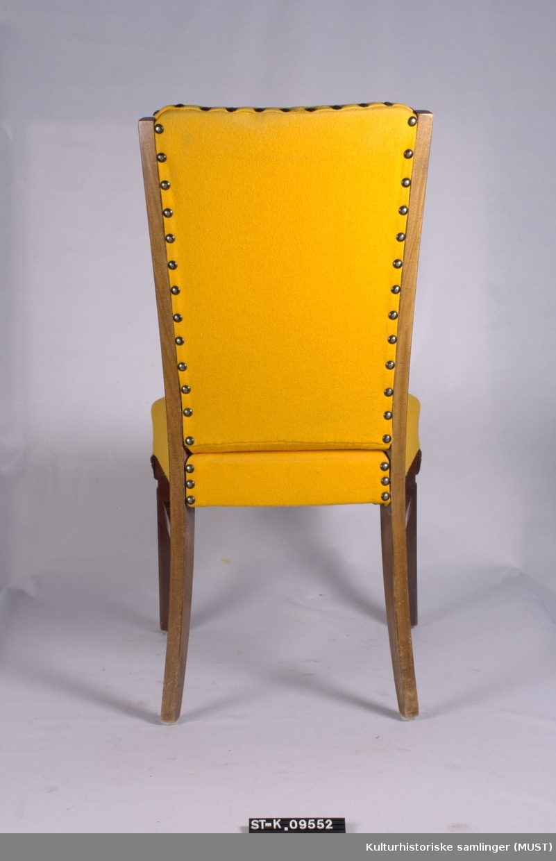 Spisestuestol med sekundært gult trekk. Stoppet rygg og sete. Frambein formet som korintiske kapitel.
