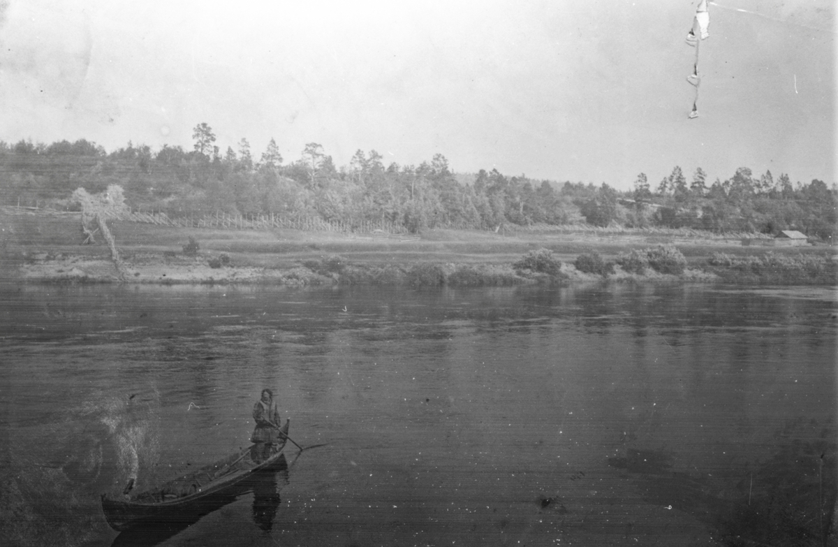 Motiv av ei kvinne i kano/ elvebåt på Karasjoks elv. På andre siden av elva er det satt opp gjerder for reindrift?