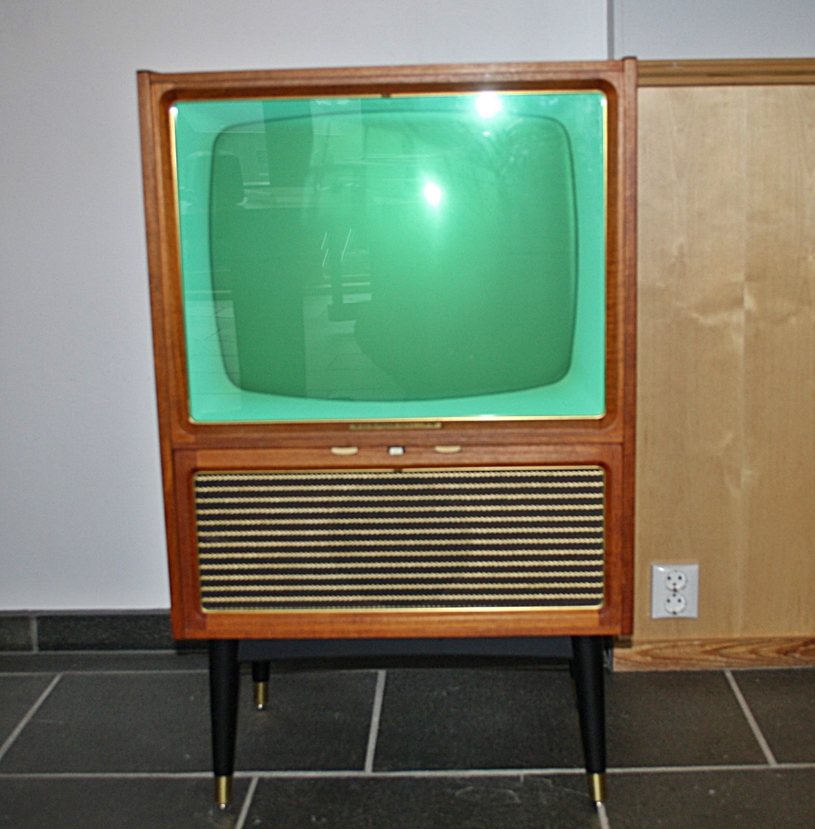 Fjernsynet står i ei kasse av tre med fire bein. Glassrute framme. Kassen er laga i finer. Ei tynn messinglist kring glassruta. Grønn skjerm. Høgtalar under skjermen.