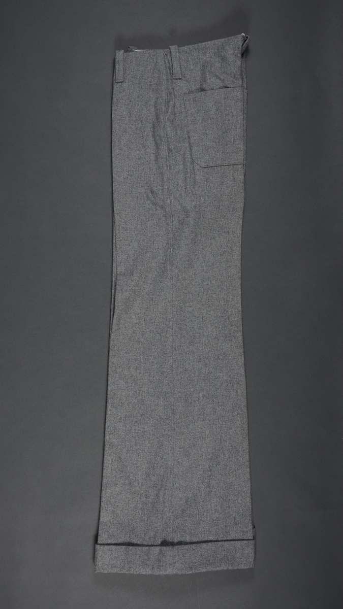 Slengbukse i grå flanell. Enkel modell uten linning men med beltestropper. Gylfåpning og knapp foran. Sleng i buksebeina, oppbrett nederst.