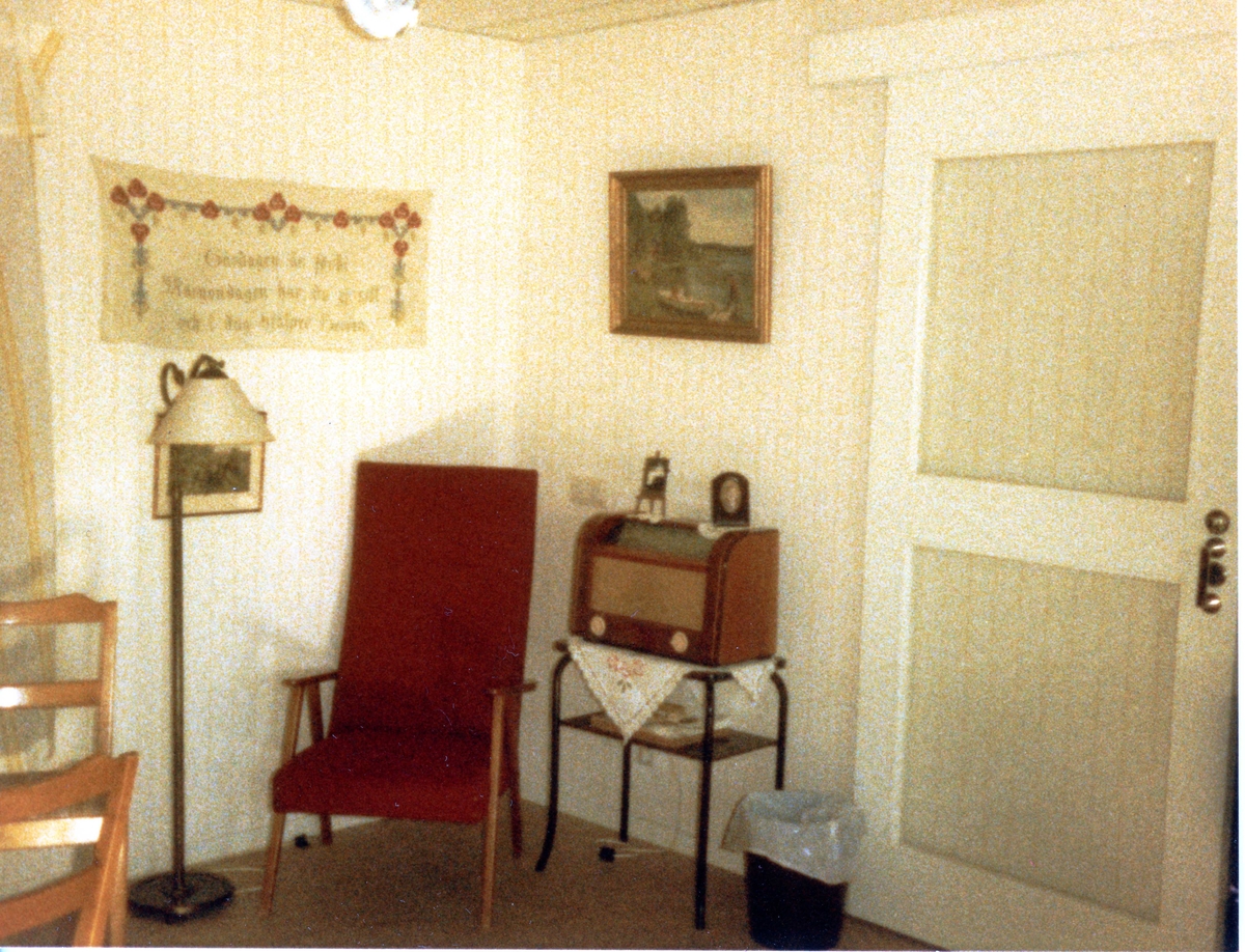 Brattåsgården vid invigningen år 1986. Utställning av föremål från gamla Brattåshemmet, gjord av Kållereds Hembygdsgille. Delar av föremålen finns nu på Mölndals stadsmuseum. Här ses en röd fåtölj, radio på ett litet bord, väggbonad och tavla.