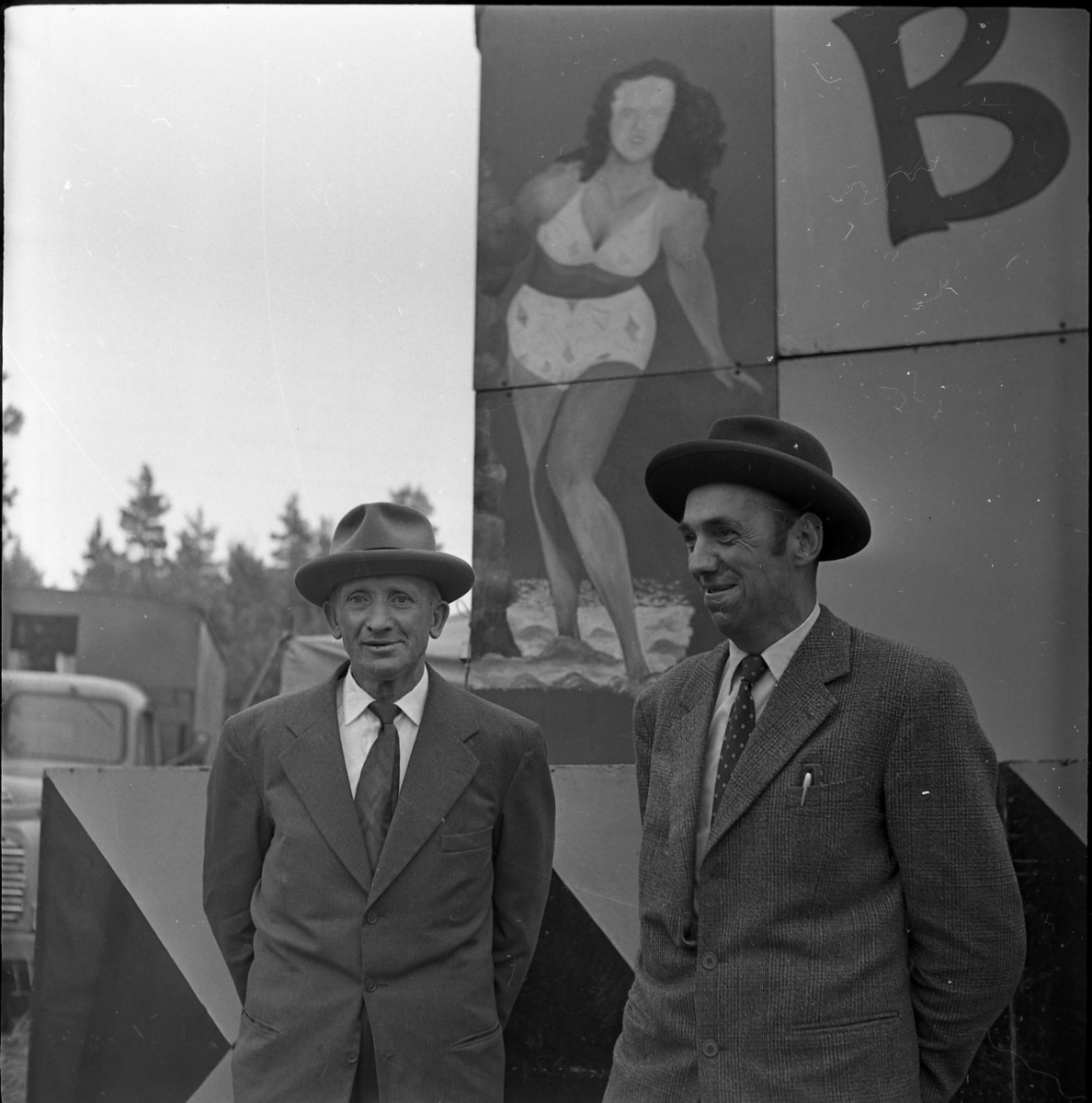 Två män med hatt står framför en reklamskylt med en lättklädd dam, typ pinuppa. Adelövs marknad. Männen är Einar Karlsson och Erik Johansson.