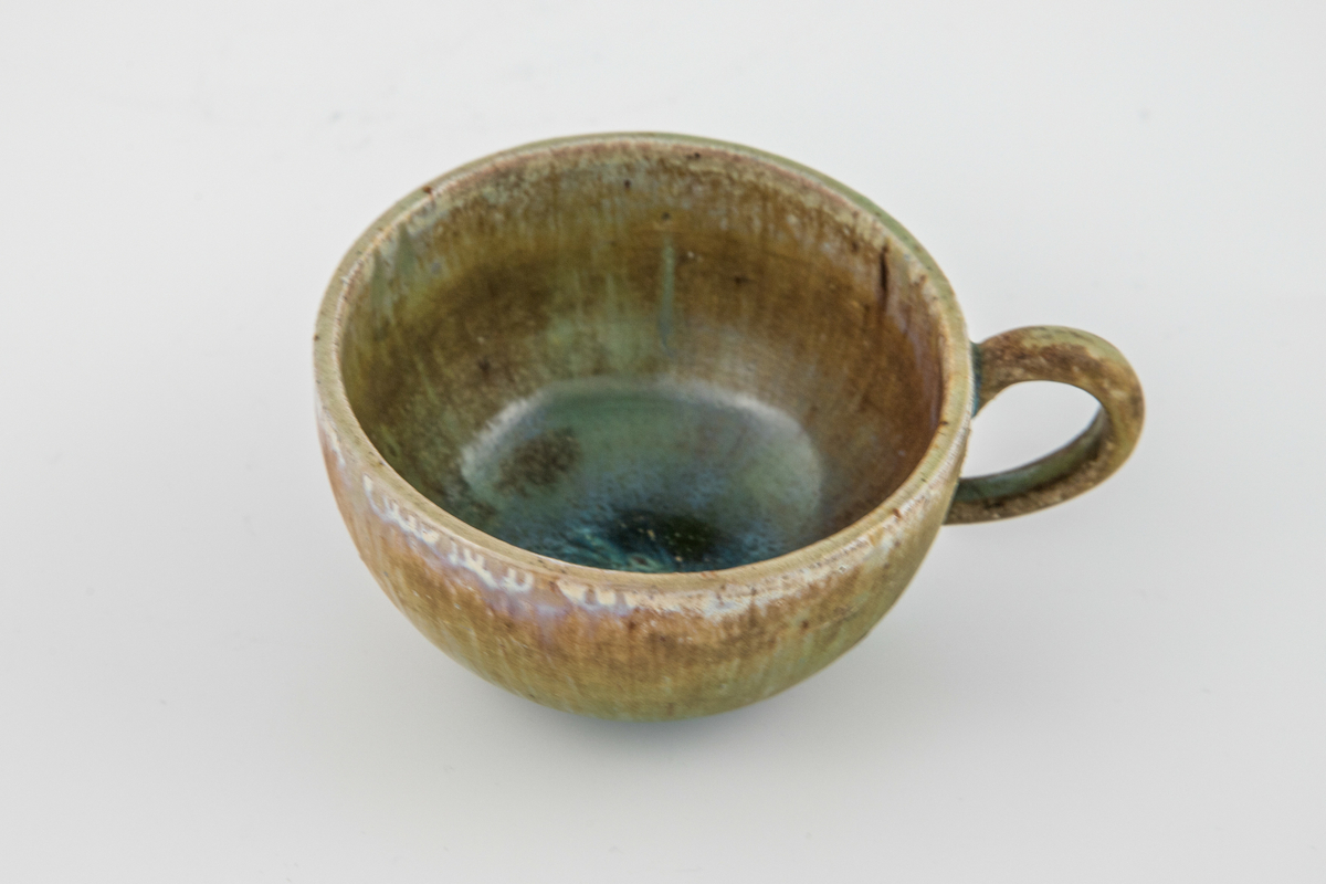 Kopp med skål i glasert steingods. Førstnevnte har påsatt hank. Både kopp og fat er dekket med en brun-grønn glasur som har rike fargevirkninger i form av blå, hvite, rosa og lilla innslag. På fatet er det små krystalliserte partier. 