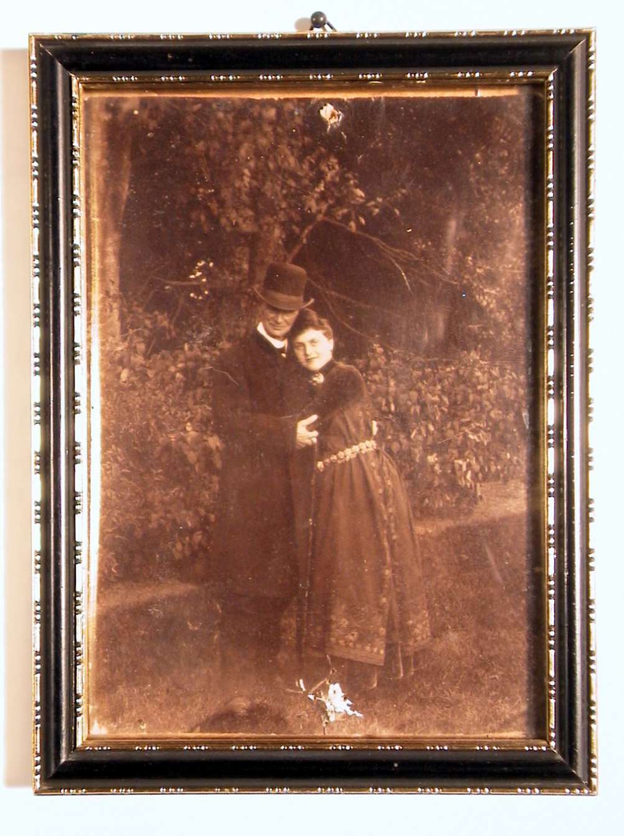 Utendørsfotografi av en mann og en kvinne med armene om hverandre. Han i mørk dress, hatt og frakk og stokk; hun i mørk bunad med sølvbelte og smykker. Hun ser direkte på betrakteren.