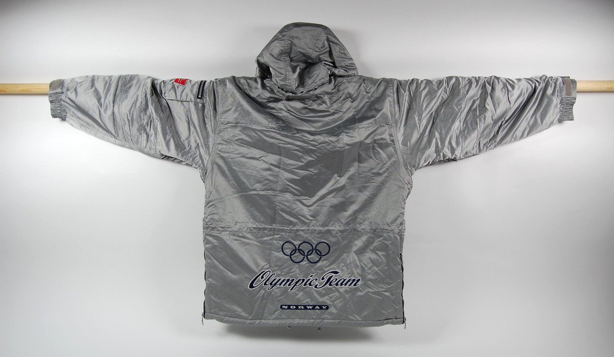 Jakke med hette. Hovedfargen er sølvgrå. Det er en blå og grå logo for Olympic Team på ryggen, og det er påpsydd et lite norsk flagg på venstre erme. Jakka har flere lommer. 