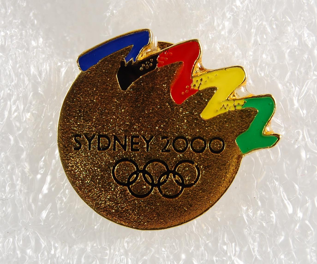 Flerfarget pins med logo for de olympiske leker i Sydney i 2000. Hoveddelen av pinsen er gullfarget. Pinsen har noe slitasje på baksiden.