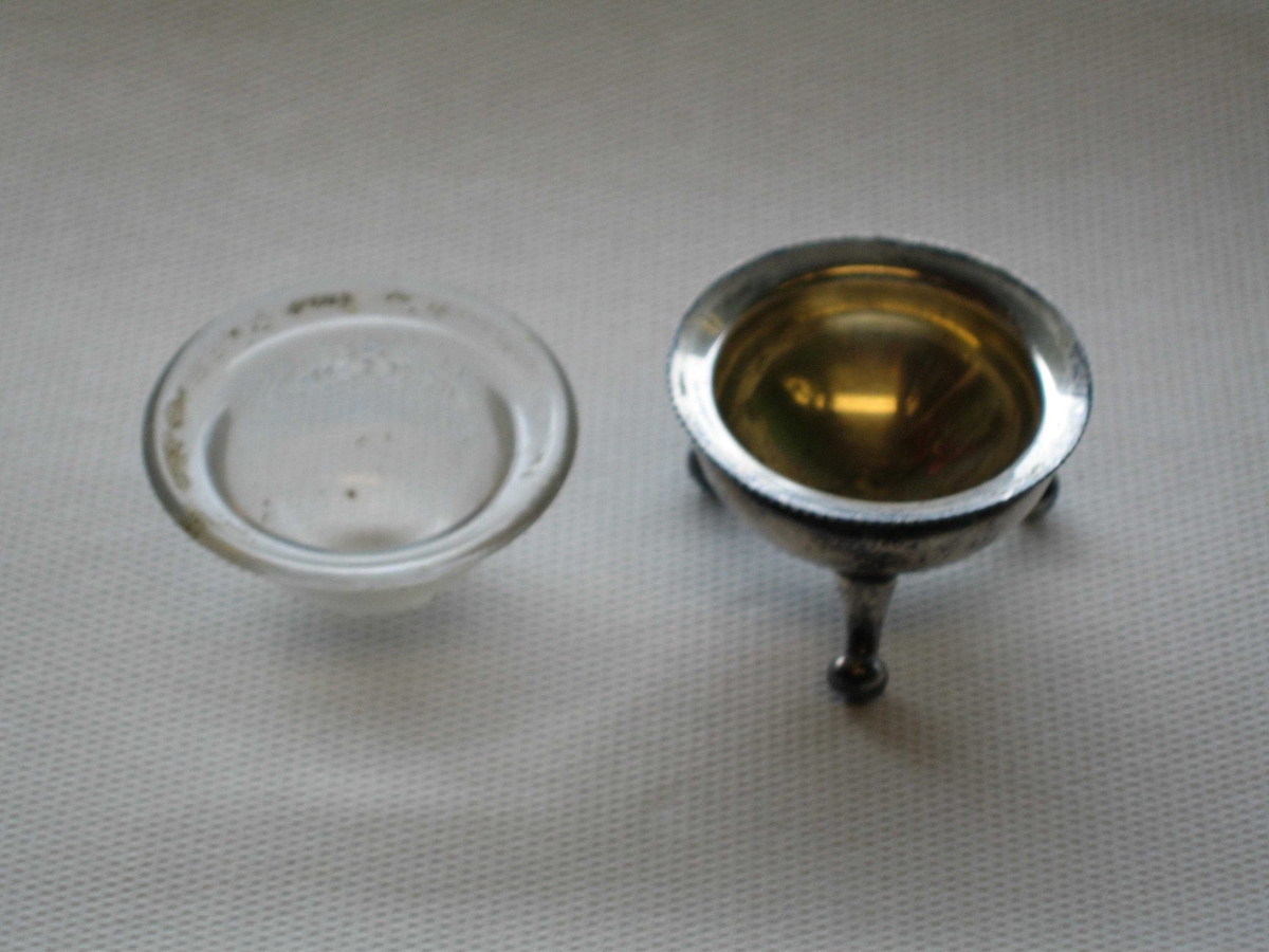 Saltkar av sølvplett og glass. Sølvfoten formet som en gryte med tre bein. Halvkuleformet glassinnlegg med utbrettet kant.