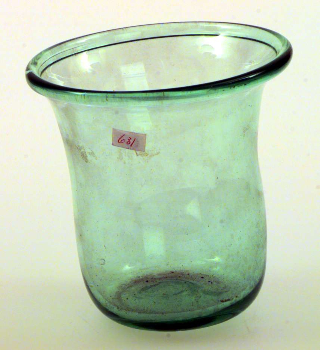 Urtepotten er av glass. Den er svakt grønn. Bunnen er ujevn. Potten er sylinderformet med utbøyet kant.