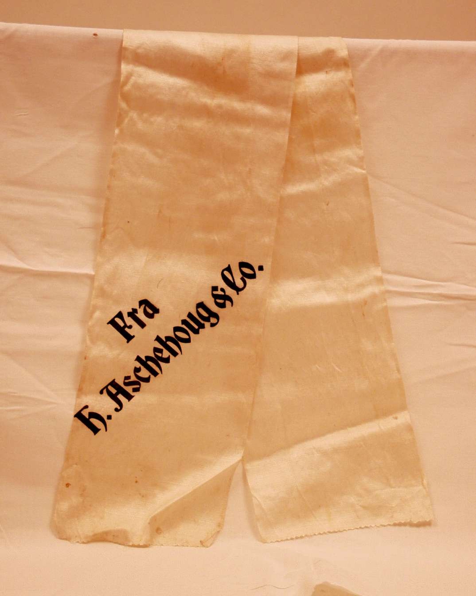 Bånd i kremfarget silke. Båndet er delt i to. På båndet står: Fra H. Aschehoug & Co.