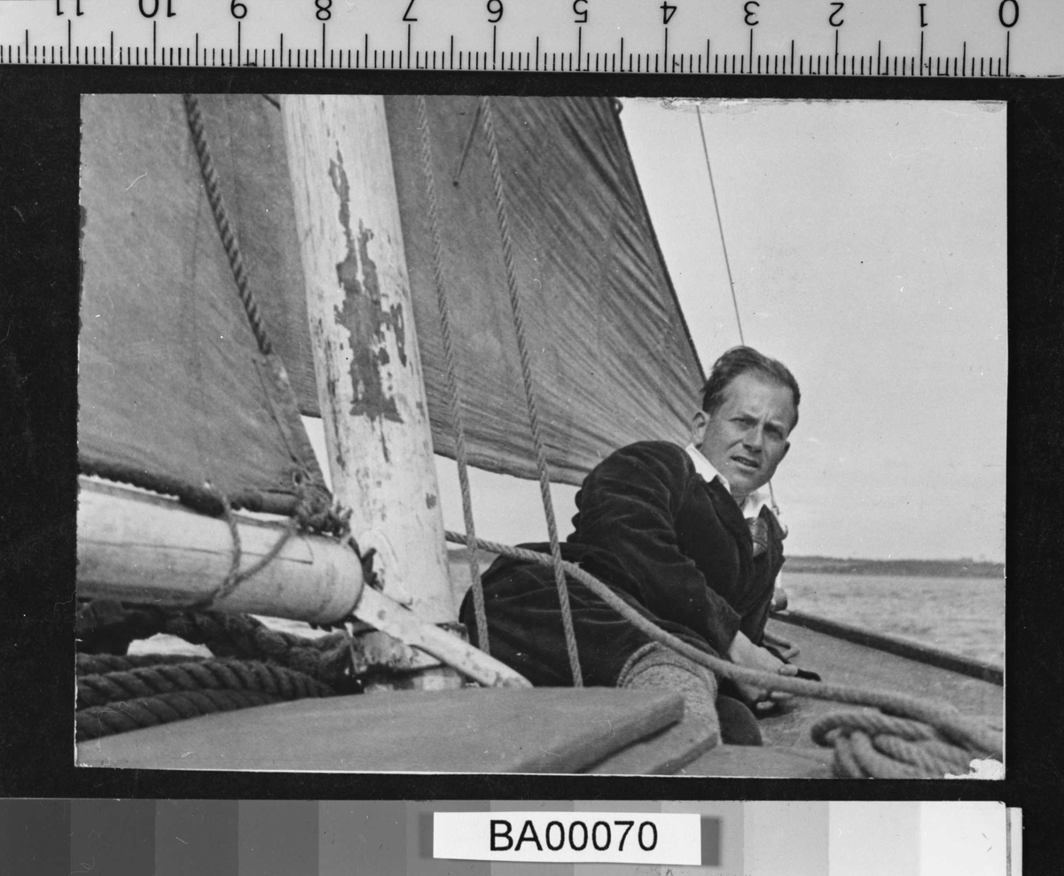 Fotografi av voksen ung mann liggende på dekket av gammeldags trebåt med kulørt seil. Han ser direkte på fotografen.