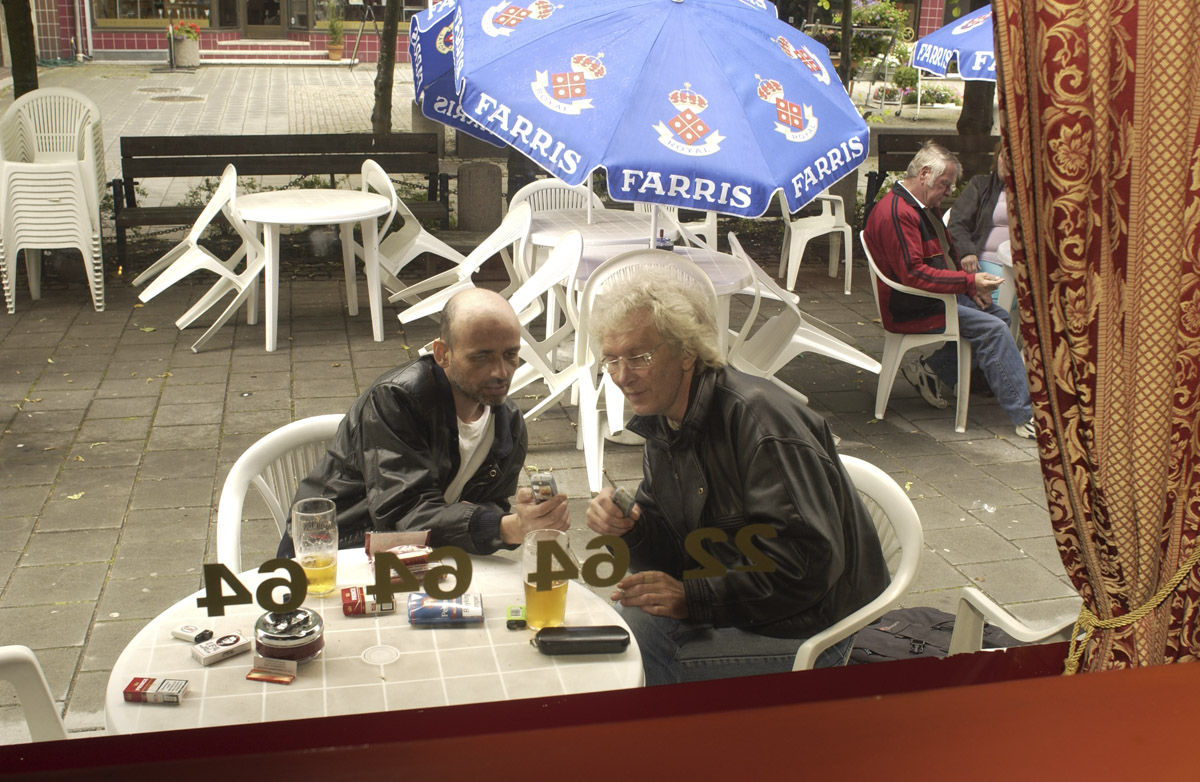 Første dagen etter innføring av røykeloven 2004. Flamenco Pizza & Pub. Parasoller og hagemøbler. To menn sitter ved et bord og røyker. Bildet er tatt innefra, gjennom vinduet.