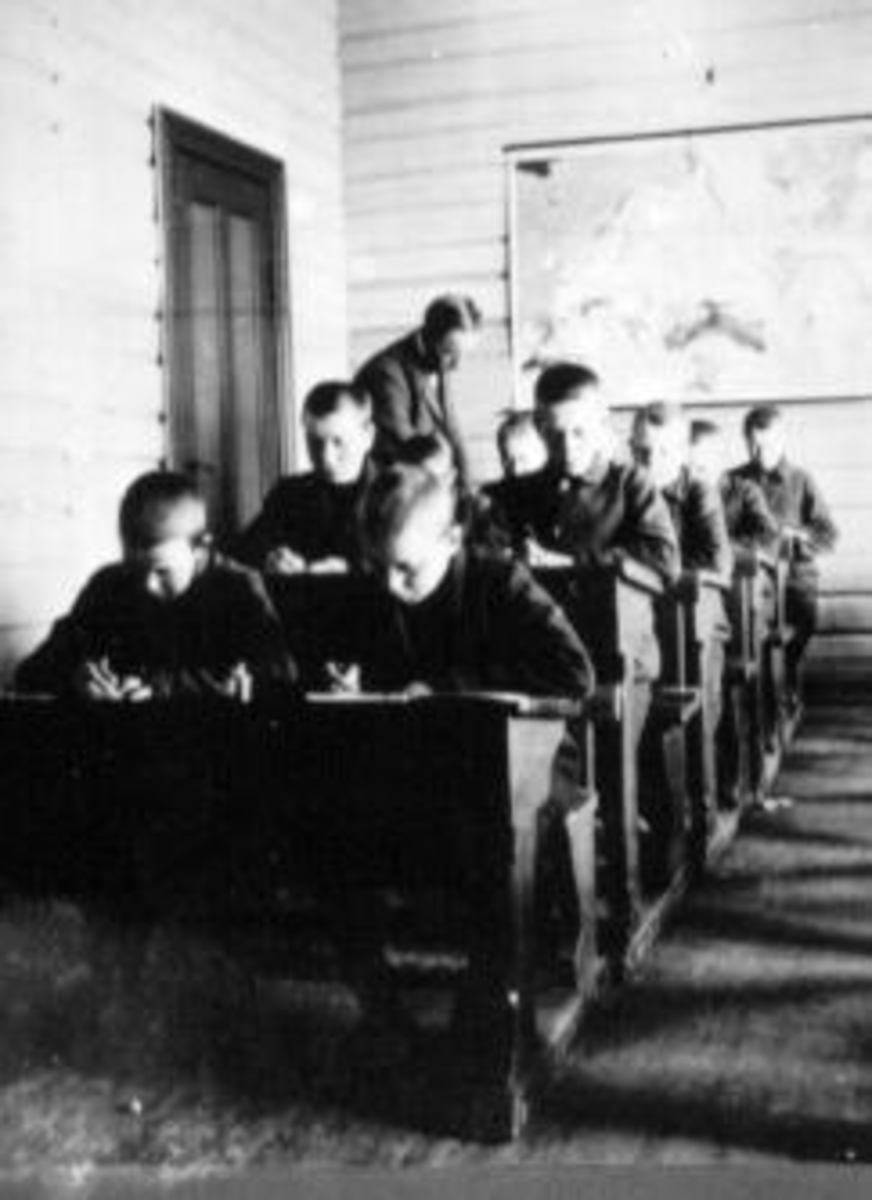 Interiør fra klasserom på Toftes Gave, Nedre Sund, Helgøya. Lærer underviser gutter som sitter ved 2-manns pulter.